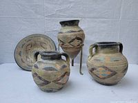 Four pottery pieces displayed including two handled jars, one footed jar on a metal stand, and one plate.