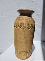 Photo of a tall beige pottery vase with vertical and geometric carved patterns, flared rim, and banded base detail in natural lighting against a white background.