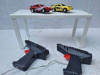 Two black handheld remote controllers with red triggers placed below a white shelf with the cars on top.