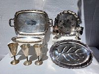 Front view of all silver plate items arranged standing and flat on a surface, showing trays, goblets, cups, and serving dish.