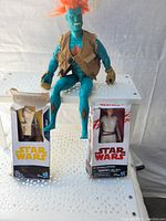 Three figures arranged on a perforated metal table: two Star Wars action figures in original boxes and one large blue creature figure without a box.