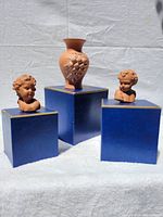 Three terra cotta pieces including an urn-style vase with a raised grape cluster on its body and two small busts of children, all displayed on blue stands.