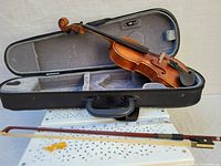 Violin inside the open case showing the violin body, fingerboard, chin rest, tailpiece, and bow lying flat inside.