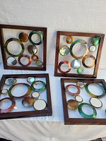 Four metal wall art pieces with circular rings and mirrors inside wooden frames on a white cloth background.