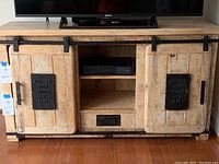 Front view of blonde wood entertainment unit with black metal barn door accents, open shelves, drawer, and sliding doors closed.