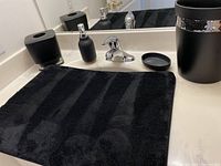 Photo showing soap pump, soap dish, tissue container, bath mat, trash can on bathroom counter, highlighting black color and mosaic tile accents on tissue container and trash can.