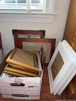 Box filled with various picture frames in gold, wood, and silver finishes. Contains smaller frames and few larger wood frames stacked behind.