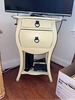 Front view of cream side table showing two drawers with ring pull handles and lower shelf with electronic accessories.