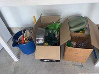 Top view of large blue container and two cardboard boxes showing various gardening tools, watering can, hoses, Miracle Gro, potting mix, and garden rocks.