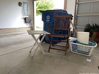 Shows green wooden folding chair, white plastic folding table, two laundry baskets (one white, one blue), and white trash can with blue wheeled recycling bin in background (not included).