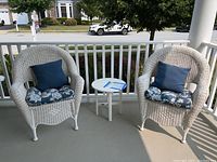 Two white wicker Hampton Bay outdoor chairs with blue floral seat cushions and navy blue pillows, flanking a small round white LL Bean outdoor table on a porch.