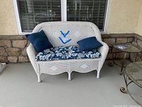 Front view showing overall white wicker love seat on porch with blue floral seat cushion and two navy blue pillows.
