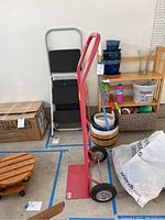 Red two-wheel hand truck standing upright on concrete floor near various household items and boxes.