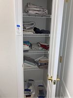 Wide view of closet shelves containing folded linens including sheets, towels, pillow cases, and floor mats. Plastic storage bins are on the floor but are not included.