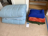 Two light blue twin-sized quilted comforters folded, placed on carpet with three throw blankets stacked next to them: royal blue, rust orange and green.