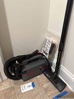 Photo of Oreck XL Pro 5 vacuum cleaner with attached hose, wand, floor nozzle, and brush attachment placed on tiled floor near corner of a room. Paper tag labeled 'Oreck Vacuum' is nearby.