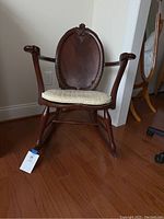 Front view of ornate rocking chair with beige seat cushion against wall and hardwood floor.