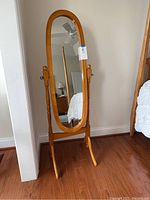 Front view of the oval cheval mirror with light wood frame and stand positioned on hardwood floor against the wall.