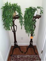 Two dark wood carved plant stands with faux plants positioned in a corner, one with an orange tag indicating repair needed.