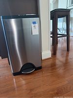 SimpleHuman stainless steel step trash can shown from front angle on wooden floor with black foot pedal visible.