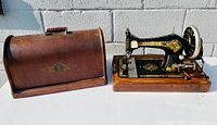 Antique Singer 28K sewing machine with wooden base next to its curved-top wooden case, both placed outside against a white brick wall.