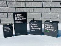 Four Cards Against Humanity boxes arranged in a row against a white brick background: First Expansion, original game, Absurd Box, Green Box.