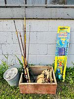 Full lot of vintage fishing rods, Supreme minnow bucket, WWII gas mask bags with fishing gear inside, two new Shakespeare fishing rod combos in original packaging, all arranged outdoors against wall.