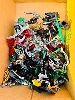 Box containing a variety of assorted LEGO Bionicle parts and partially assembled figures in multiple colors including red, black, grey, green, yellow, and silver.