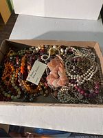 Box with assorted beaded necklaces including faux pearls, gold-tone chains, and peach carved necklaces.