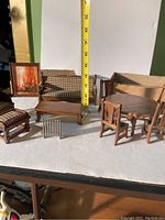 Wooden doll house furniture including sofa, hassock, coffee table, stove/fireplace, dining table, and chairs shown on white surface with measuring tape for scale.