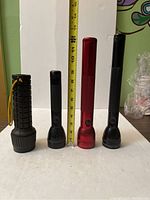 Four flashlights of various sizes standing upright next to a measuring tape showing heights approximately 6 to 11 inches.