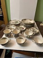 Full set of Johnson Bros The Friendly Village patterned dishes displayed with cups, saucers, plates, bowls, sugar bowl, and creamer on white cloth background
