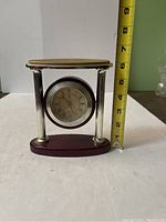 Table clock with round clock face held between two silver-tone fluted pillar columns. Wooden oval base and top platform in reddish-brown wood visible. Measuring tape shows about 6.5 inches tall.
