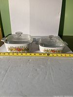 Two CorningWare LeChalet "Spice of Life" dishes with glass lids placed side by side with measuring tape showing length of larger dish approx. 10 inches.