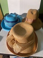 Photo showing three hats - light brown felt hat with pink ribbon, tan woven straw hat with netted bow, bright blue fabric hat with striped ribbon and scarf.