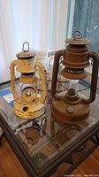 Two old oil lanterns on a glass tabletop, one yellow and one rusty brown