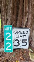 Two street signs leaning against a tree: Speed Limit 35 (white with black text) and Mile 22 (green with white text). Shows some dirt and natural outdoor wear.