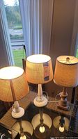 Three table lamps on a table near window, turned on showing illuminated fabric shades, two white lamp bases with some scratches, one light brown lamp base with shade damage