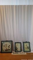 Front view of three framed Asian bird artworks, all featuring feather designs, arranged side by side on floor.