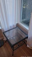 Square glass top metal end table positioned in a corner, showing clear glass surface and metal frame with decorative scrollwork lower shelf.