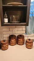 Set of 4 ceramic kitchen jars labeled Tea, Flour, Sugar, Coffee with wood barrel design and round black knob lids on kitchen counter.
