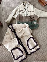 Two wool cardigans laid out on carpet, showcasing Norwegian cardigan with sheep and nature design and English cardigan with purple trim pockets.