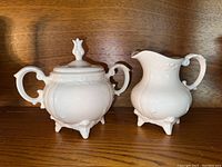 Two photos showing a white ceramic hand-painted sugar bowl with lid and matching creamer pitcher with ornate handles and feet, placed on a wooden shelf.