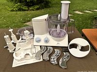 Photo showing Braun Multiquick 3 K650 food processor base with clear processing bowl, plastic pusher, multiple stainless steel slicing and shredding blades, two vertical blade holders and instruction booklet on a table