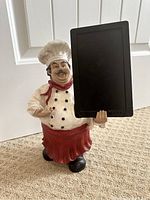Front view showing chef figurine holding black chalkboard, with detailed chef uniform and smiling expression.