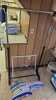Full view of black rolling clothing rack with horizontal bar and white flat mesh drying rack sitting behind it, and the 13 covered hangers lying in front on carpeted floor.