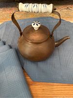 Copper teapot with porcelain handle and knob, placed on blue placemats and alongside folded blue napkins on a wooden surface