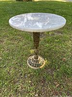Full view of the round marble top table resting on grass, showing its heavy marble surface and ornate brass base.