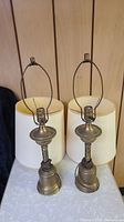 Pair of brass tone metal table lamps with original off-white fabric shades, shown side by side.