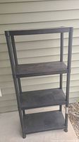 Full view of the black plastic 4-shelf storage unit against an exterior wall, showing height and shelf spacing.
